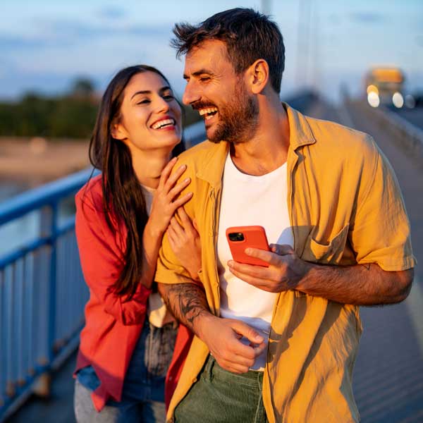 Laughing couple on a bridge with a phone at sunset.